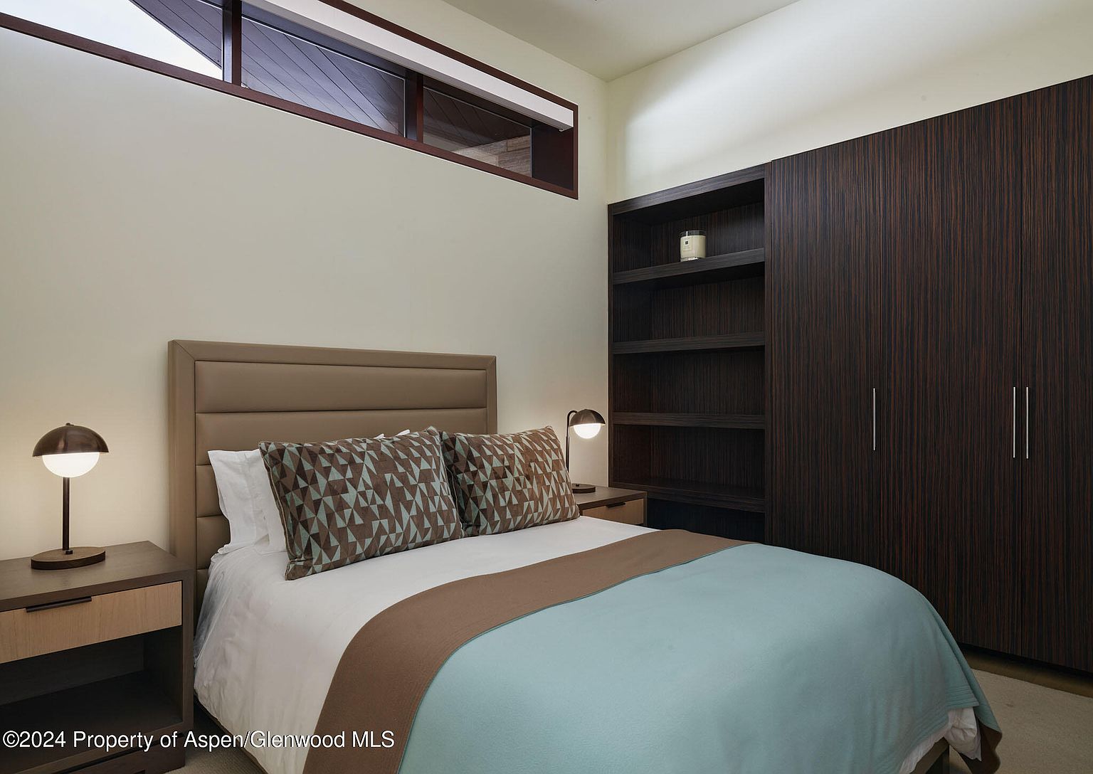 This is a cozy guest bedroom featuring a queen-sized bed with a light blue coverlet and brown accent pillows. A modern nightstand with a lamp sits on either side of the bed. A dark wood built-in wardrobe and shelving unit provides ample storage. A clerestory window provides natural light.