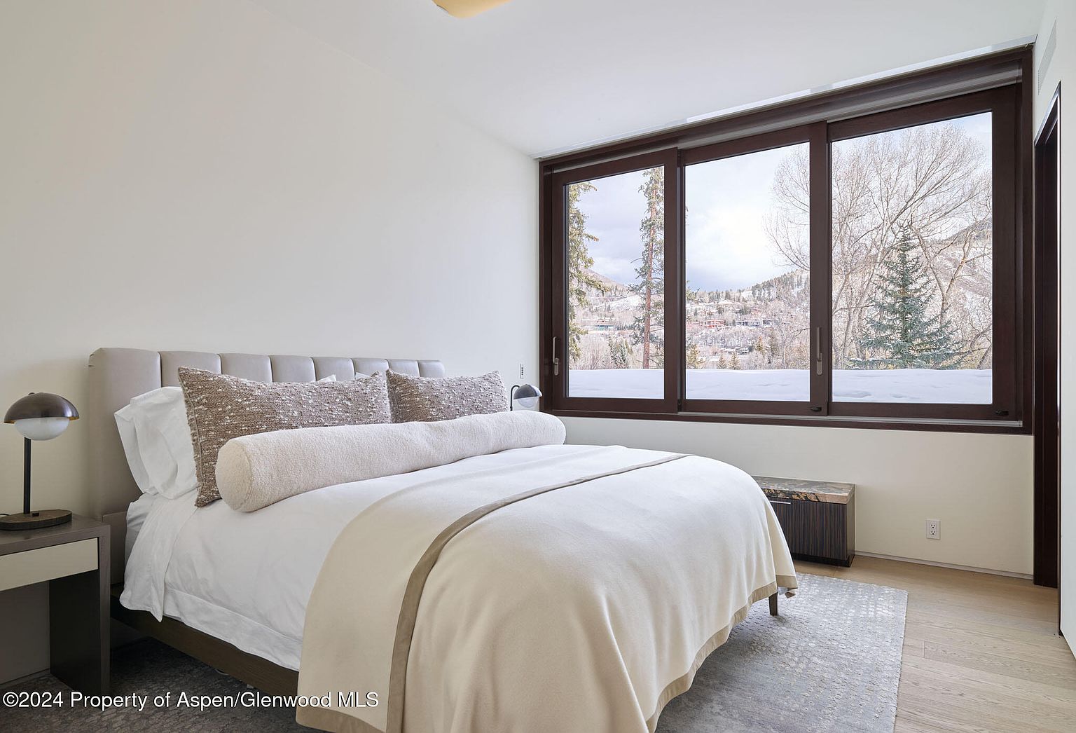This is a well-lit primary bedroom featuring a large bed with neutral-toned bedding and pillows. A dark wood-framed window offers a view of a snowy landscape, and a small side table with a lamp sits beside the bed. The room has a minimalist and serene aesthetic, perfect for relaxation.
