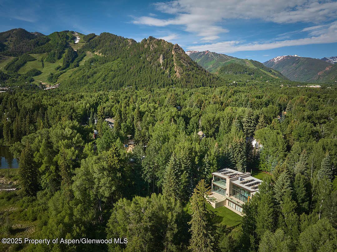 This aerial view showcases a modern home nestled amidst lush greenery, with mountains visible in the background. The house features a contemporary design with clean lines and a flat roof, blending seamlessly with the natural surroundings. The landscape is dominated by dense trees, creating a sense of privacy and tranquility.