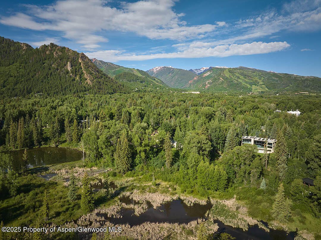 This aerial view showcases a property nestled amidst lush greenery and mountains. A modern house is visible among the trees, with a pond and marshy area in the foreground. The landscape provides a sense of privacy and natural beauty, highlighting the property's serene setting.