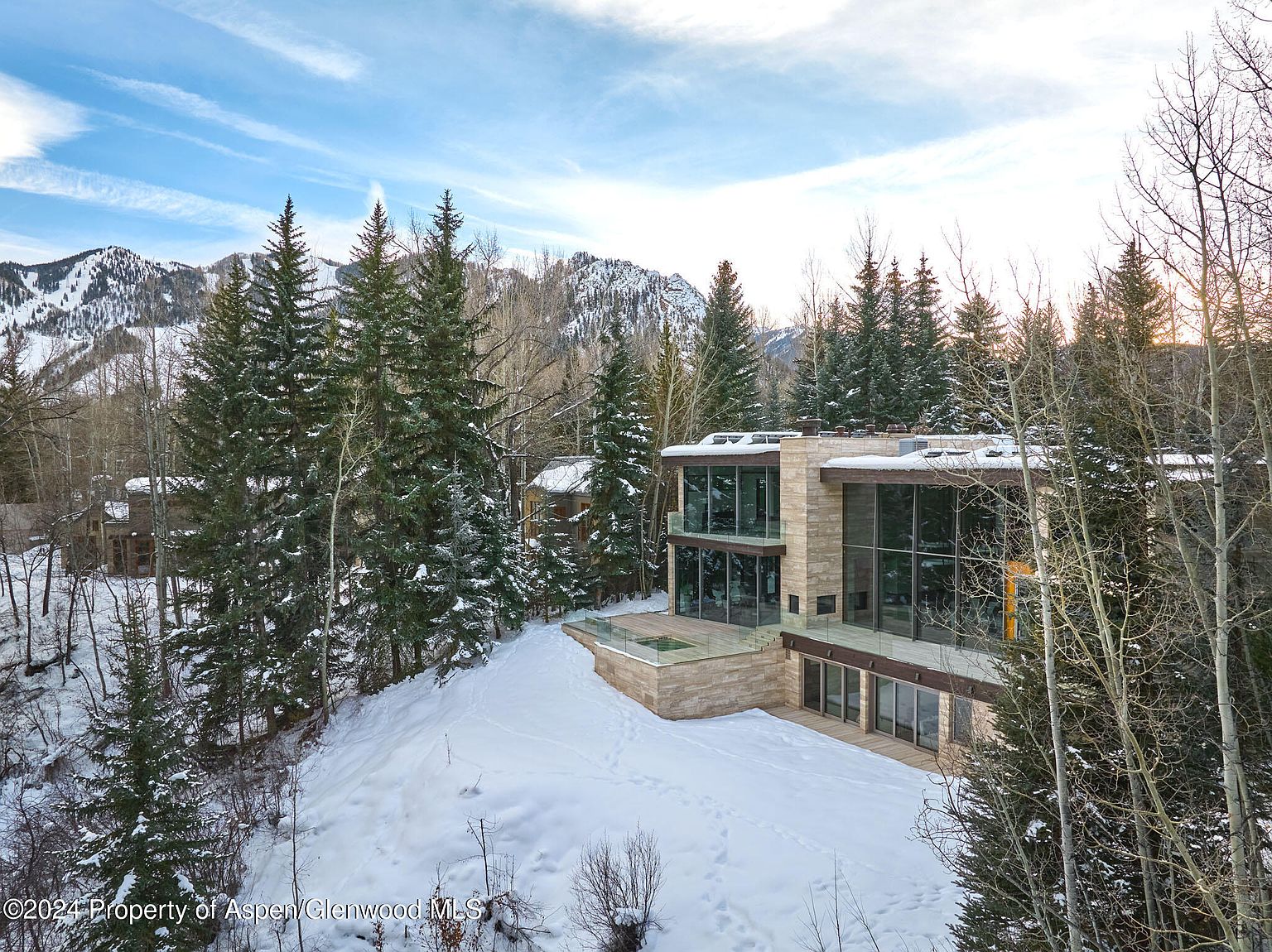 This image showcases the rear exterior of a modern home, emphasizing its architectural design and integration with the snowy, wooded landscape. The house features large windows and a multi-level deck, offering stunning views of the surrounding mountains and trees. The snow-covered yard adds a serene and picturesque quality to the property.