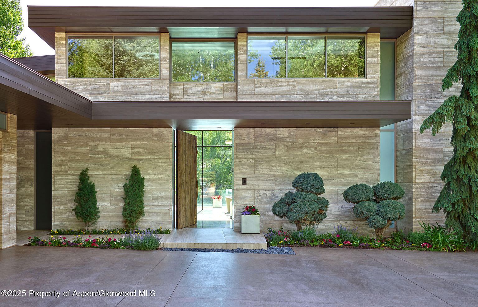 This is a front exterior shot of a modern home, emphasizing the entryway. The facade is clad in horizontally laid stone, complemented by dark brown trim and large windows reflecting the surrounding greenery. The open wooden door invites entry, flanked by manicured greenery and colorful flowerbeds, creating a welcoming and sophisticated curb appeal.