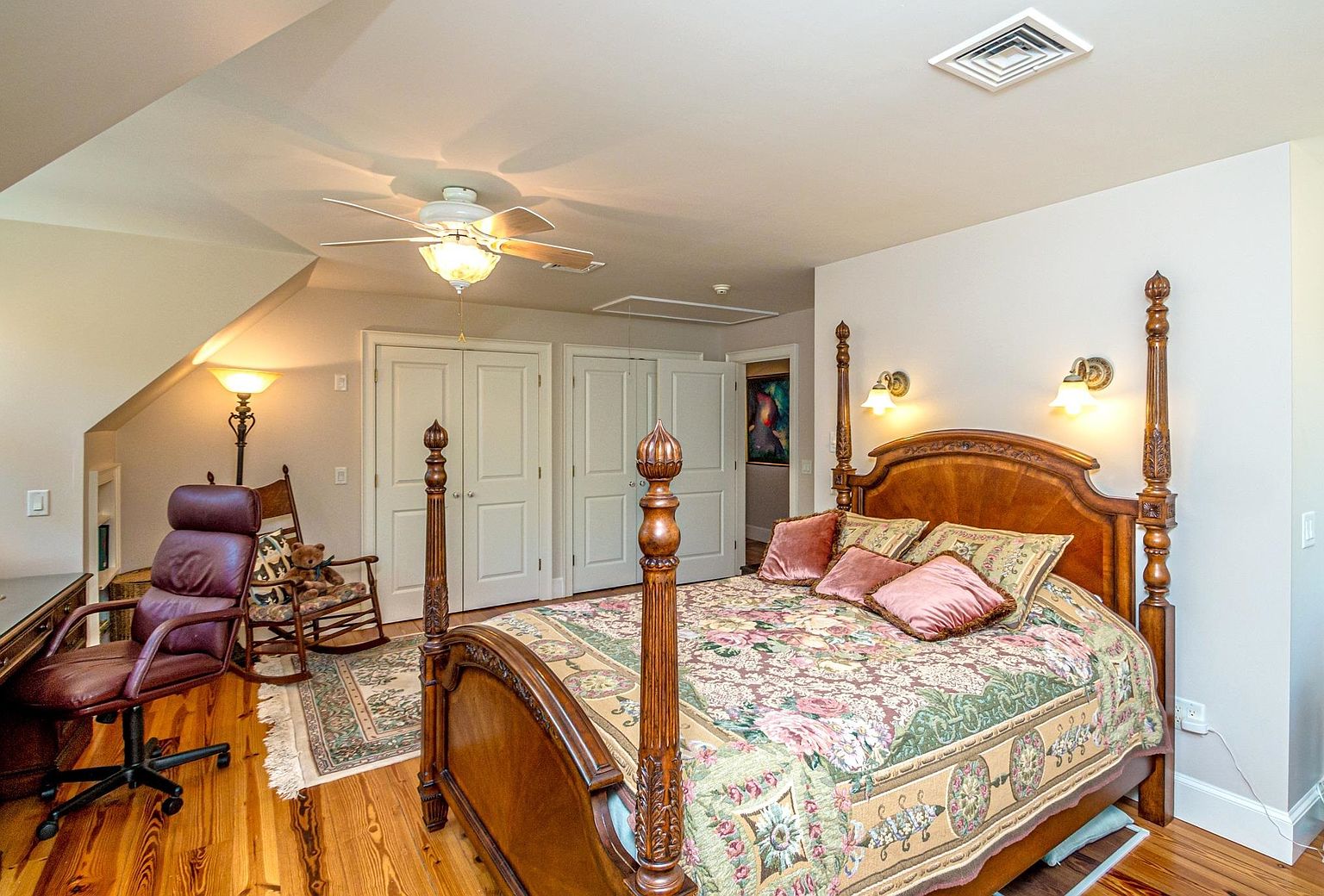 This primary bedroom features a traditional four-poster wooden bed as the centerpiece, dressed in a floral patterned quilt with decorative pillows. To the left, a dedicated workspace includes a leather office chair and a desk, complemented by a nearby rocking chair and a floor lamp. The room is finished with warm wood flooring, neutral walls, and multiple doors leading to closet spaces, creating a cozy and functional living environment.