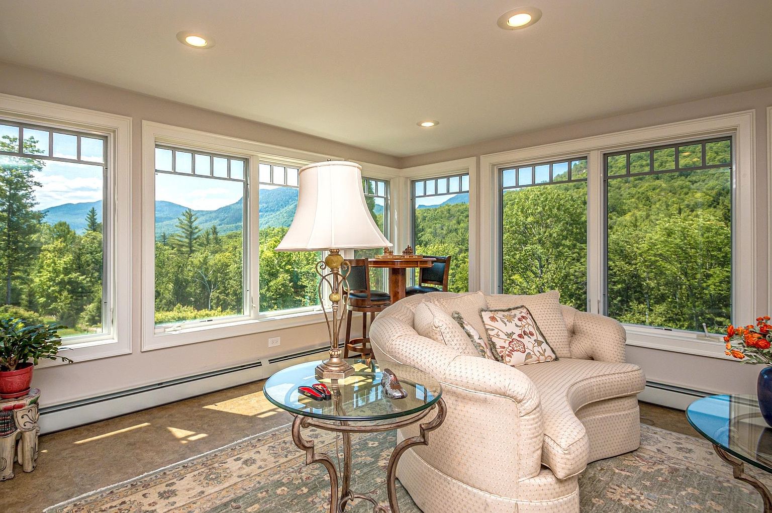 This bright and airy sunroom features a curved, upholstered armchair positioned to take advantage of the expansive, panoramic views of the surrounding mountains and lush forest. A glass-topped side table with a decorative lamp sits beside the chair, while a small bistro table and chairs are tucked into the corner near the large windows. The space is defined by its neutral color palette, patterned area rug, and abundant natural light, creating a serene and inviting atmosphere.