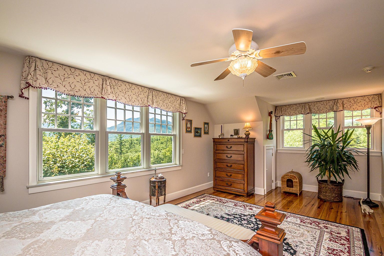 This spacious primary bedroom features warm wood flooring, a large patterned area rug, and a classic wooden bed frame. Large windows with decorative valances provide ample natural light and scenic mountain views, while a ceiling fan with a decorative light fixture centers the room. A tall wooden dresser and a potted plant add character to the space, creating a cozy and inviting atmosphere.