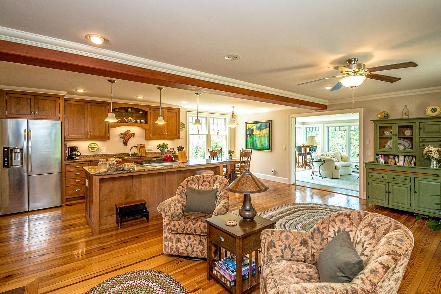 This open-concept kitchen and living area features warm wood cabinetry, a central island with a cooktop, and rich hardwood flooring throughout. The space is illuminated by recessed lighting and decorative pendants, creating a cozy, inviting atmosphere that flows seamlessly into an adjacent sunroom. A green hutch adds a touch of character, while the comfortable seating area in the foreground provides a perfect spot for relaxation.