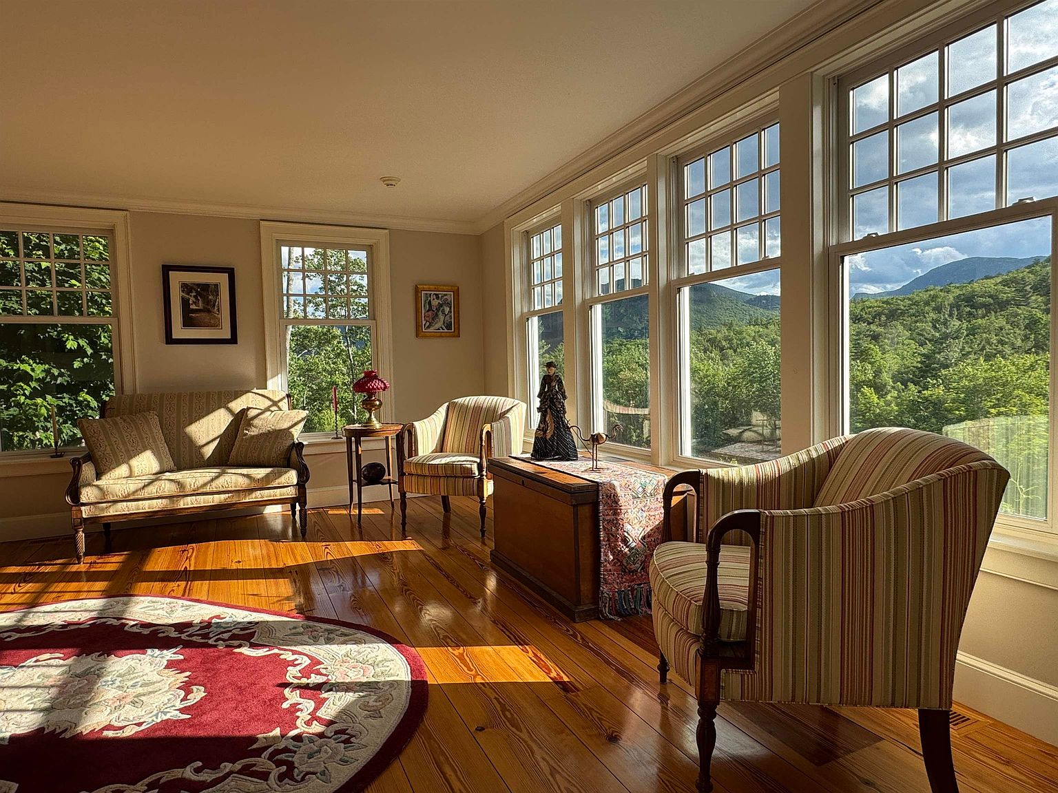 This sun-drenched sunroom features warm wood flooring and a classic, traditional aesthetic with striped upholstered seating. A series of large windows lines the exterior wall, framing expansive views of the lush green mountains and forest beyond. The space is furnished with a sofa, two armchairs, and a wooden chest, creating a cozy and inviting atmosphere perfect for relaxation.