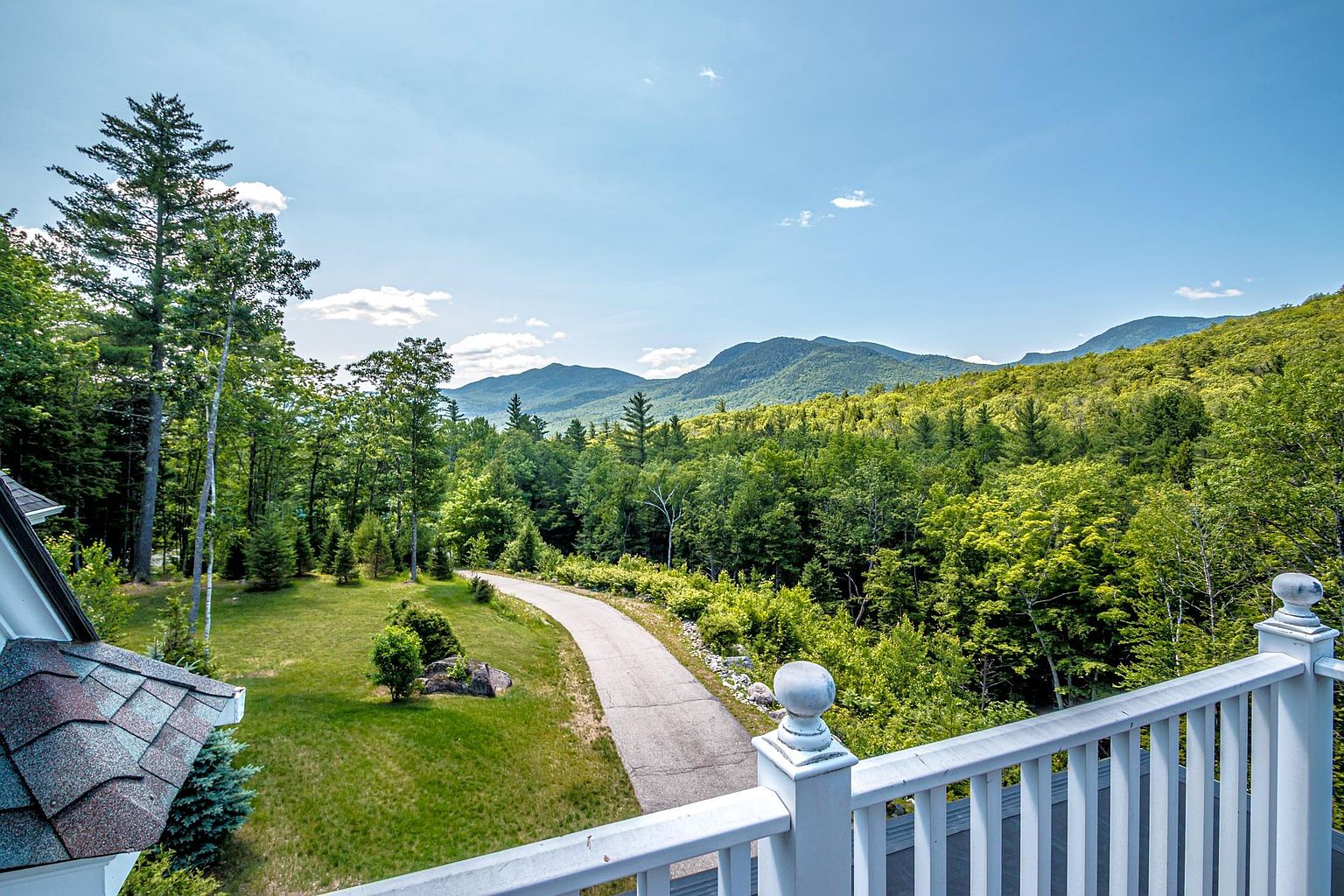 This elevated perspective from a white-railed balcony captures a sweeping, panoramic view of a lush, green mountain range under a bright blue sky. Below, a paved driveway winds through a well-maintained lawn and dense forest, highlighting the property's secluded and scenic setting. The vantage point emphasizes the expansive natural landscape and the peaceful, private atmosphere of the home.