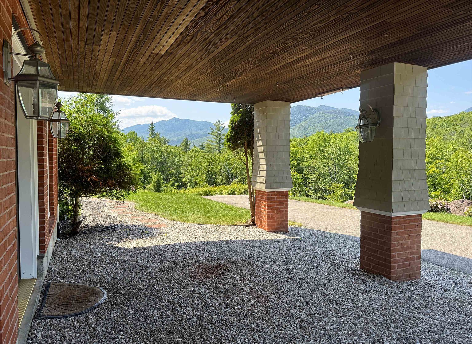 This covered entryway features a rustic wood-paneled ceiling and substantial brick-based columns topped with shingled siding. The space opens onto a gravel driveway and provides a picturesque, unobstructed view of rolling mountains and lush green trees. The perspective is from under the porch, looking outward toward the scenic landscape.