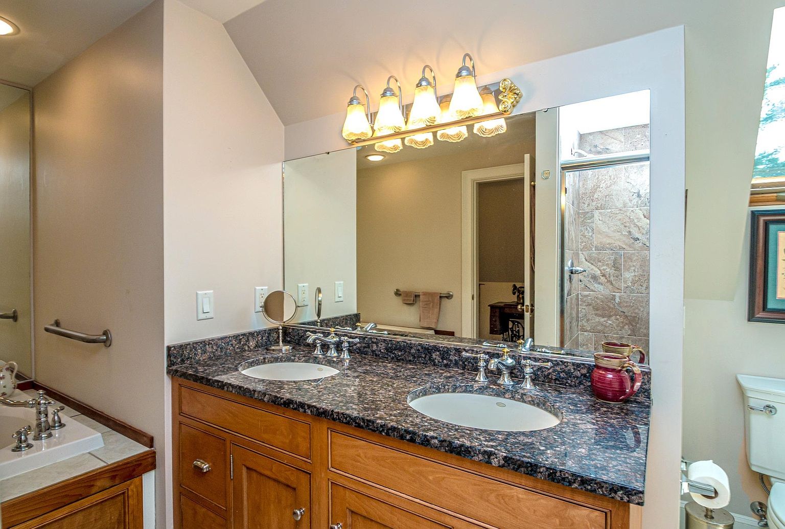 This primary bathroom features a dual-sink vanity with a dark, speckled stone countertop and warm-toned wood cabinetry. A large, wide mirror spans the wall above the vanity, illuminated by a multi-light fixture, while a tiled shower enclosure is visible in the reflection. The space includes a toilet to the right and a neutral color palette, creating a functional and traditional aesthetic.