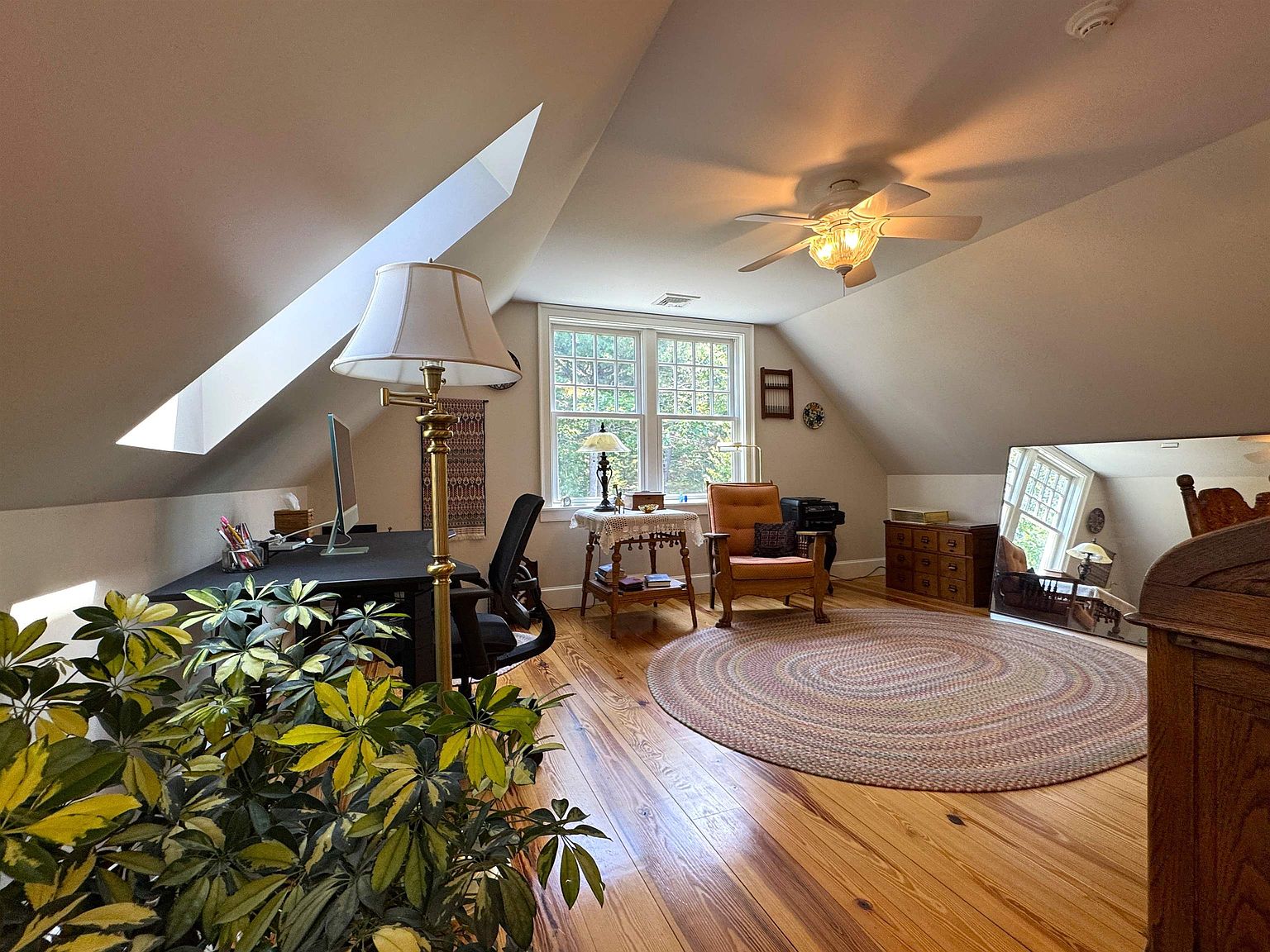 This attic office space features warm wood flooring and a sloped ceiling with a skylight, creating a cozy and inviting atmosphere. The room is furnished with a dark desk, a comfortable armchair, and a small side table, all arranged around a large, round braided rug. A large mirror reflects the windowed area, enhancing the sense of space and light in this charming, multi-functional room.