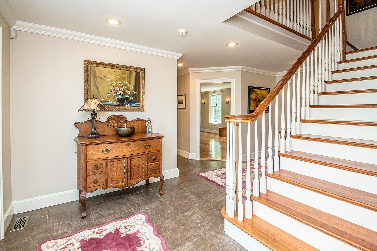 This inviting foyer features a classic staircase with white balusters and warm-toned wood treads, leading to an upper level. To the left, an antique-style wooden sideboard sits against a neutral wall, topped with a decorative lamp and a framed floral painting. The space is finished with large-format floor tiles and a patterned area rug, creating a warm and welcoming transition area that opens into a bright, adjacent room.