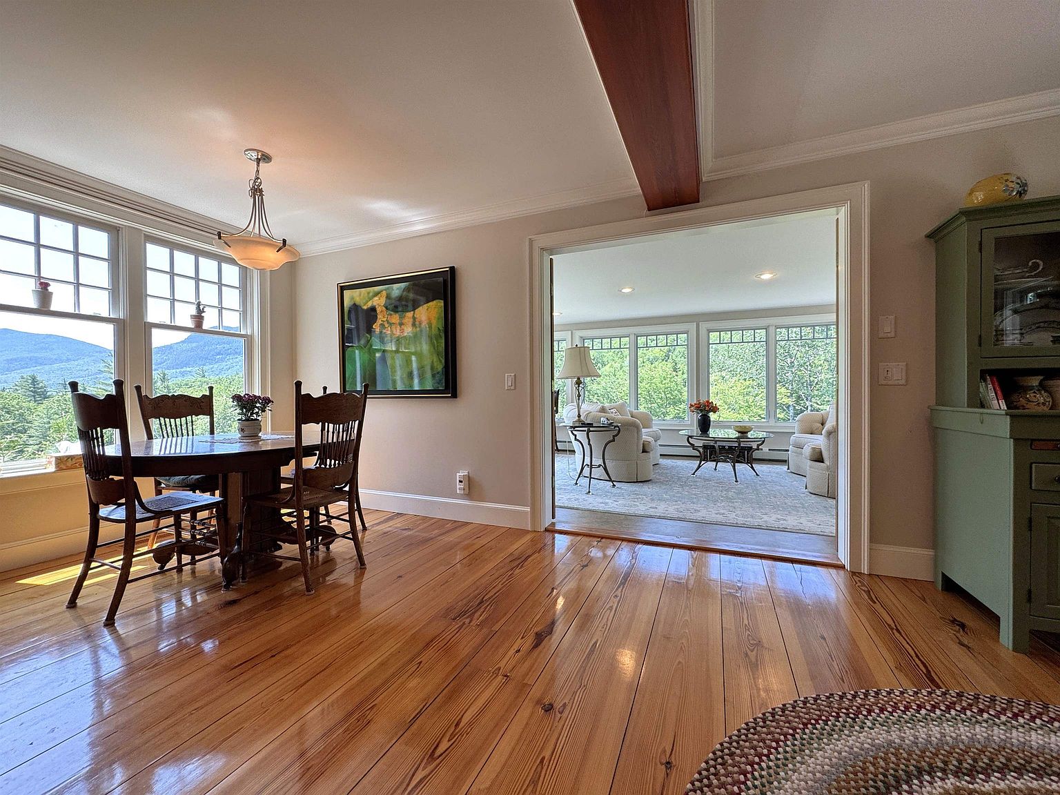 This bright dining room features warm, polished wood flooring and a classic wooden dining set positioned near large windows that offer a scenic view of the surrounding landscape. An open doorway leads into an adjacent sun-drenched living area, creating a seamless flow between spaces. The room is accented by a decorative green cabinet and a large piece of abstract wall art, contributing to a welcoming and traditional atmosphere.
