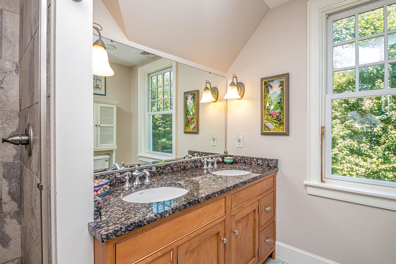 This bathroom features a double-sink vanity with a dark, speckled stone countertop and light wood cabinetry. A large, wall-to-wall mirror reflects the space, including two wall-mounted light fixtures and framed artwork, while a window to the right provides natural light. The room has a clean, functional layout with a neutral color palette and a glimpse of a tiled shower area on the left.
