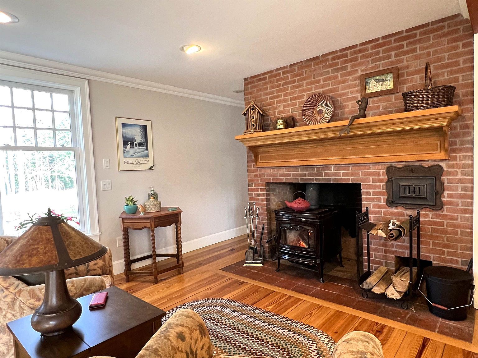 This cozy living room features a prominent brick fireplace with a wood-burning stove and a substantial wooden mantel. The space is anchored by warm-toned wood flooring and a braided area rug, complemented by comfortable patterned armchairs and a small side table. A large window allows natural light to fill the room, creating a welcoming and traditional atmosphere.