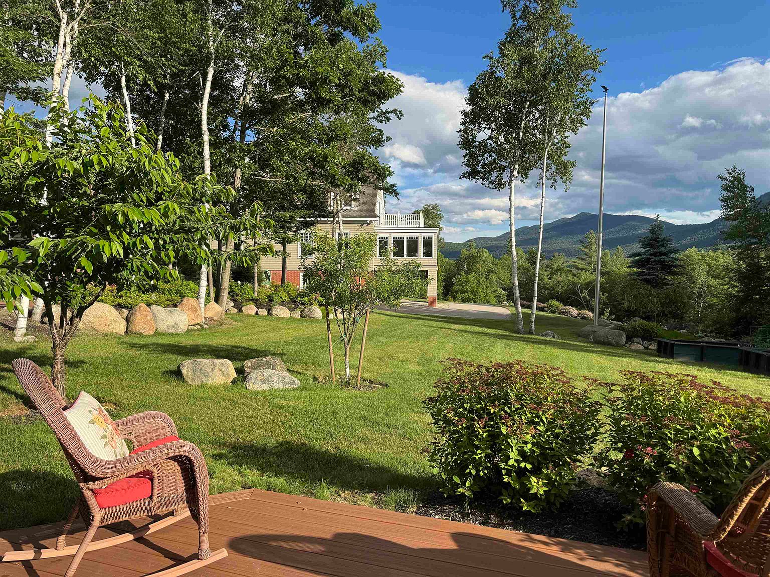 This scenic backyard view features a well-manicured lawn leading toward a multi-story home with a prominent deck and sunroom. The foreground includes a comfortable wicker rocking chair on a wooden deck, while the background showcases lush trees and rolling mountain vistas under a bright blue sky. The perspective is from a relaxing outdoor seating area, emphasizing the peaceful and expansive nature of the property.