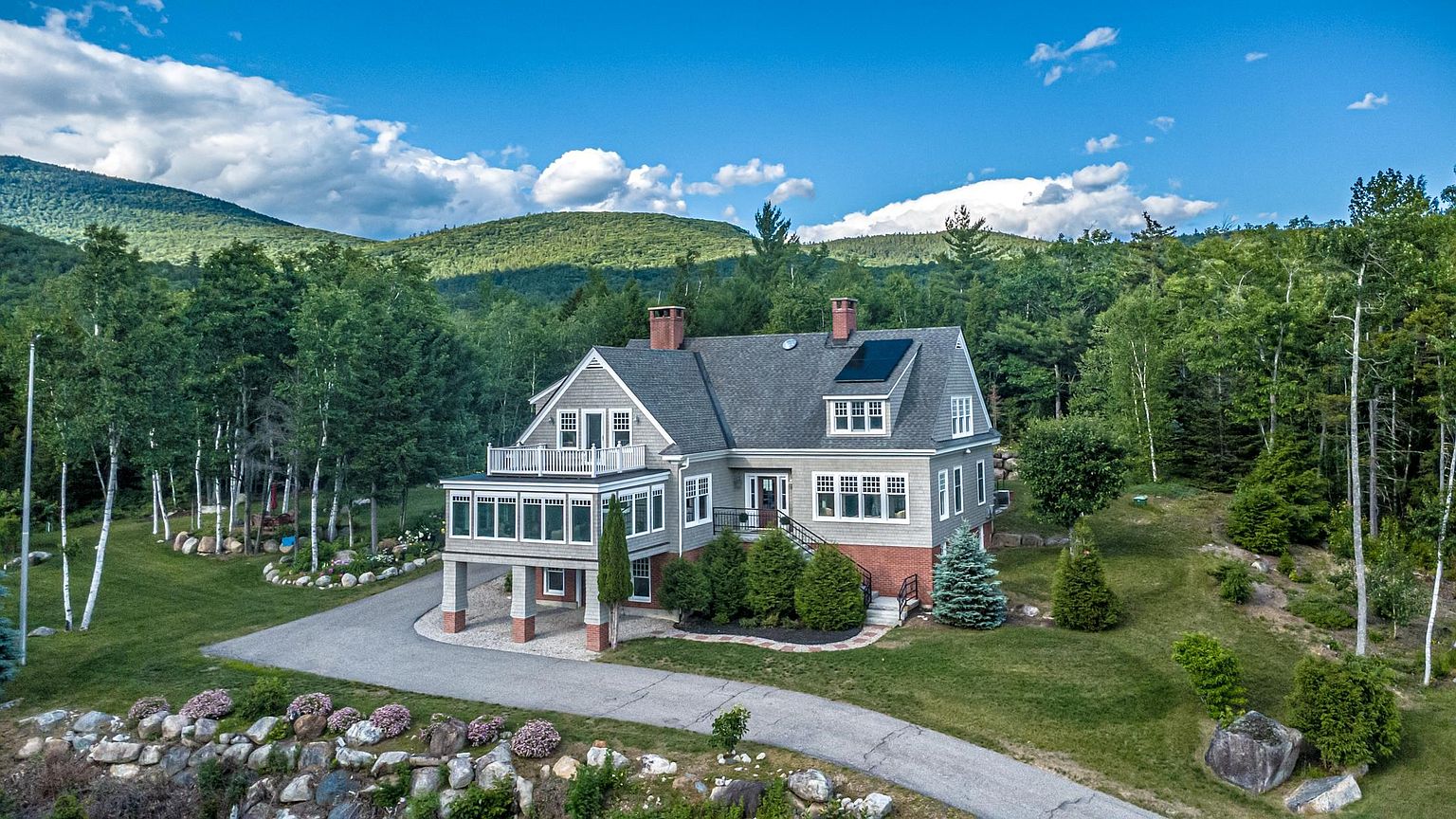 This aerial perspective captures a charming, multi-level shingle-style home nestled against a lush, forested mountain backdrop. The residence features a prominent sunroom with a balcony above, a brick-accented foundation, and a paved driveway that curves through the landscaped grounds. The setting is serene and private, showcasing a blend of traditional architectural elements and natural beauty.