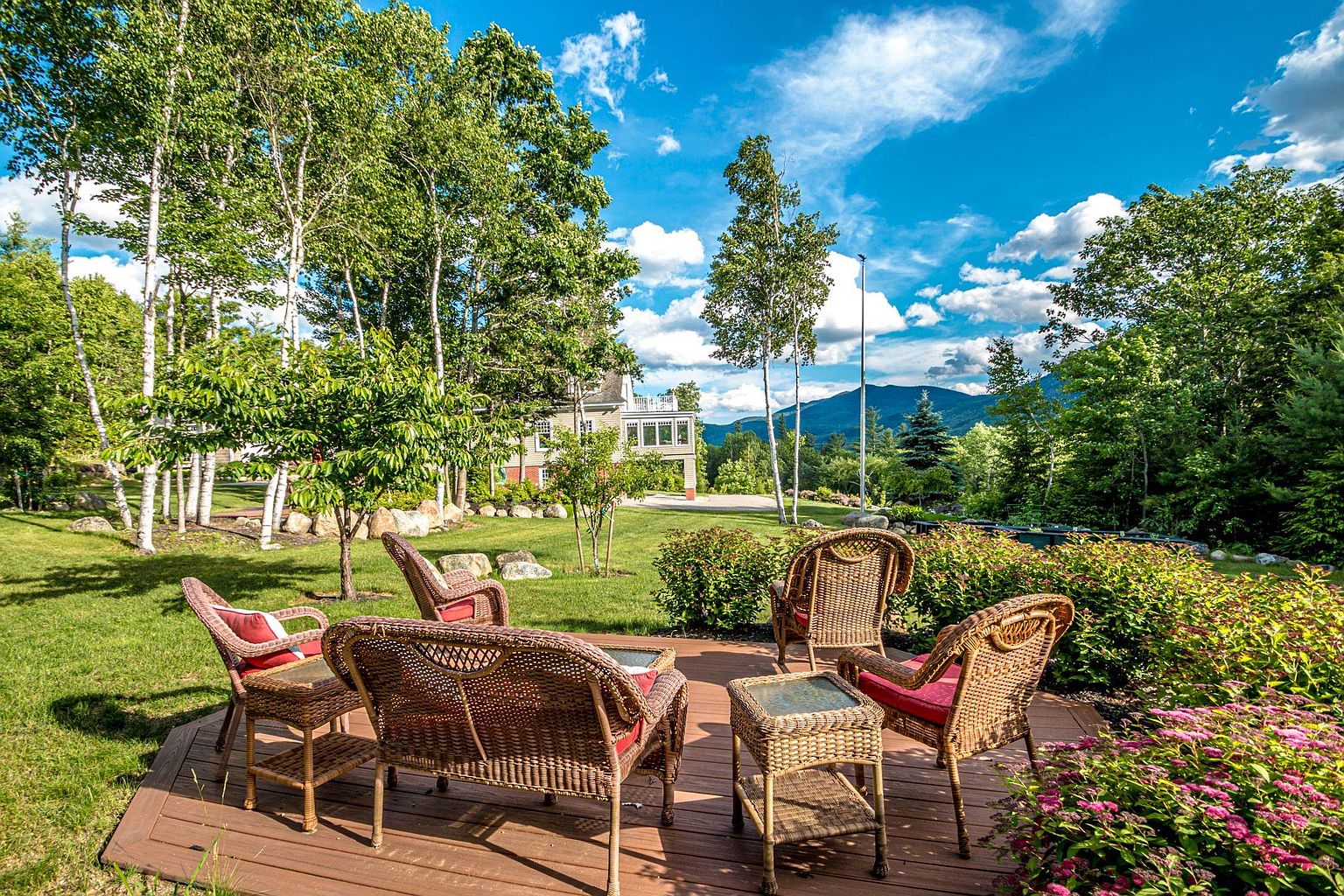 This inviting outdoor living space features a composite deck furnished with a wicker seating set, including chairs and a small table with red cushions. The deck overlooks a lush, expansive lawn surrounded by mature trees and vibrant flowering bushes, with a clear view of distant mountains under a bright blue sky. The perspective is from the deck looking out toward the property, capturing a serene and picturesque backyard setting.