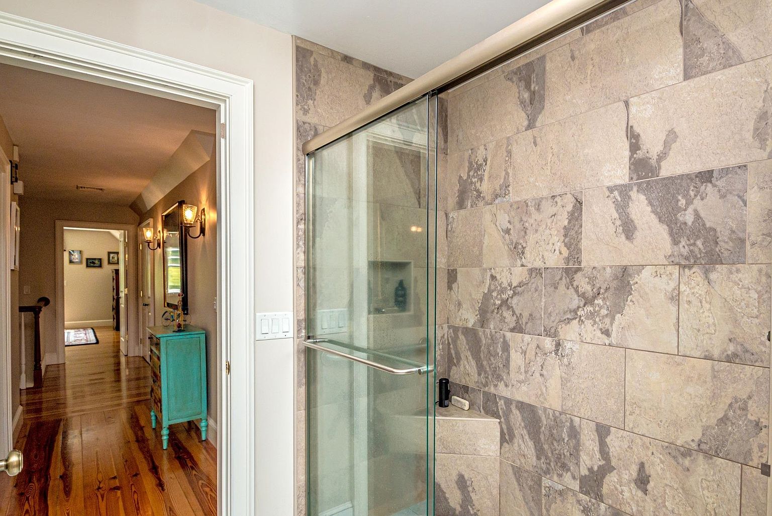This view captures a modern bathroom shower enclosure featuring large, textured stone tiles in a neutral palette. The glass shower door is framed in a dark metal, and the perspective looks through the doorway into a hallway with warm-toned wood flooring and a vibrant teal accent cabinet. The transition between the tiled bathroom space and the adjacent hallway creates a sense of depth and flow within the home.