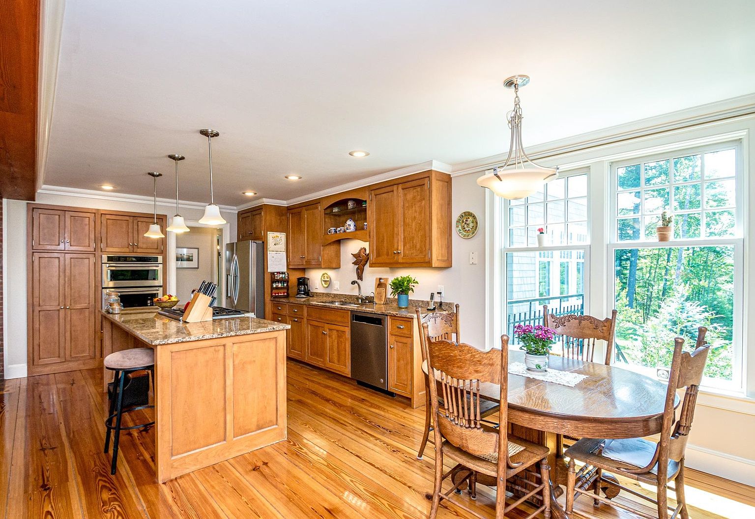This warm and inviting kitchen features medium-toned wood cabinetry, granite countertops, and a central island with bar seating. A dining area is positioned adjacent to the kitchen, illuminated by natural light streaming through large windows that overlook the surrounding trees. The space is finished with polished wood flooring, creating a cohesive and traditional aesthetic perfect for family gatherings.