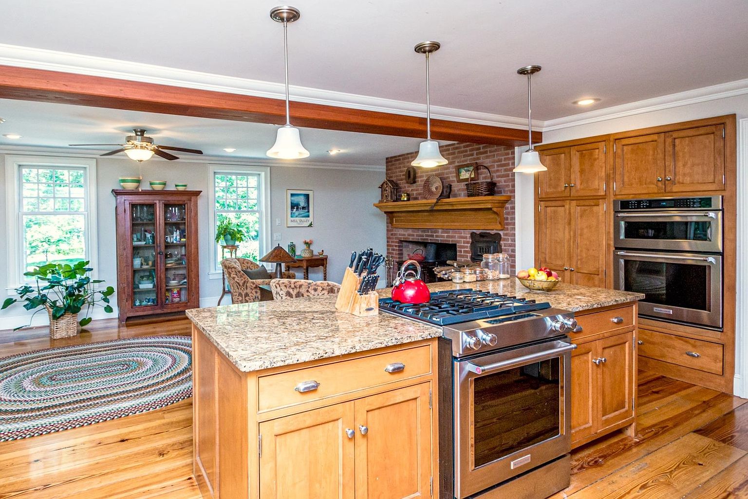 This open-concept kitchen features light wood cabinetry, a large central island with a granite countertop, and stainless steel appliances, including a double wall oven and a gas range. The space flows into an adjacent living area, which is anchored by a brick fireplace with a wooden mantel and a cozy seating arrangement. Warm lighting from pendant fixtures and natural light from the windows create an inviting, traditional atmosphere.