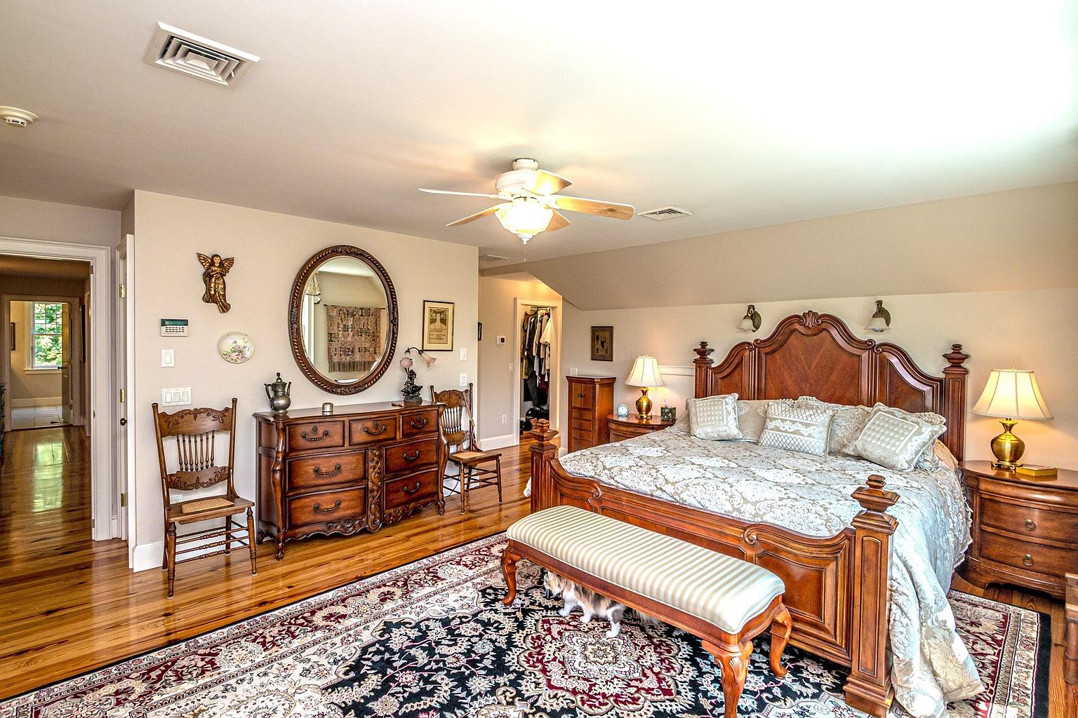 This spacious primary bedroom features a large, ornate wooden bed frame as the centerpiece, complemented by a matching dresser and nightstands. The room is decorated with a traditional aesthetic, including a patterned area rug, a striped bench at the foot of the bed, and a large oval mirror. A ceiling fan provides overhead lighting, while warm lamps on the nightstands create a cozy atmosphere, and a glimpse into an adjacent hallway and walk-in closet adds depth to the space.