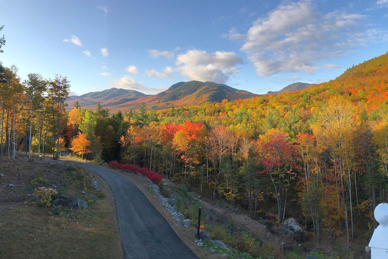This elevated perspective captures a scenic driveway winding through a vibrant, autumn-foliage landscape. The view showcases a dense forest canopy painted in rich shades of orange, red, and yellow, set against a backdrop of rolling mountains under a clear blue sky. The composition emphasizes the property's secluded, natural setting and expansive mountain vistas.