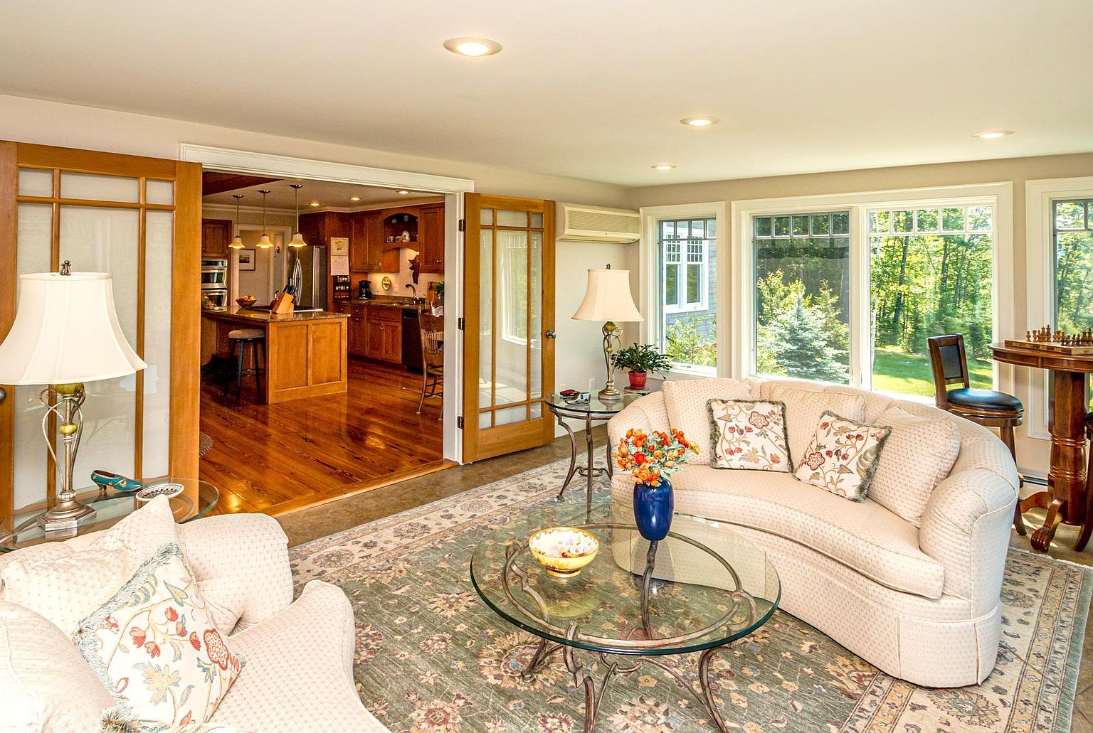 This bright and inviting living room features a curved sofa with patterned throw pillows, a glass-topped coffee table, and a large, intricate area rug. The space flows seamlessly into an adjacent kitchen through wide, multi-pane glass doors, creating an open and airy atmosphere. Large windows along the right wall provide ample natural light and views of the surrounding greenery, while a small game table sits in the corner.