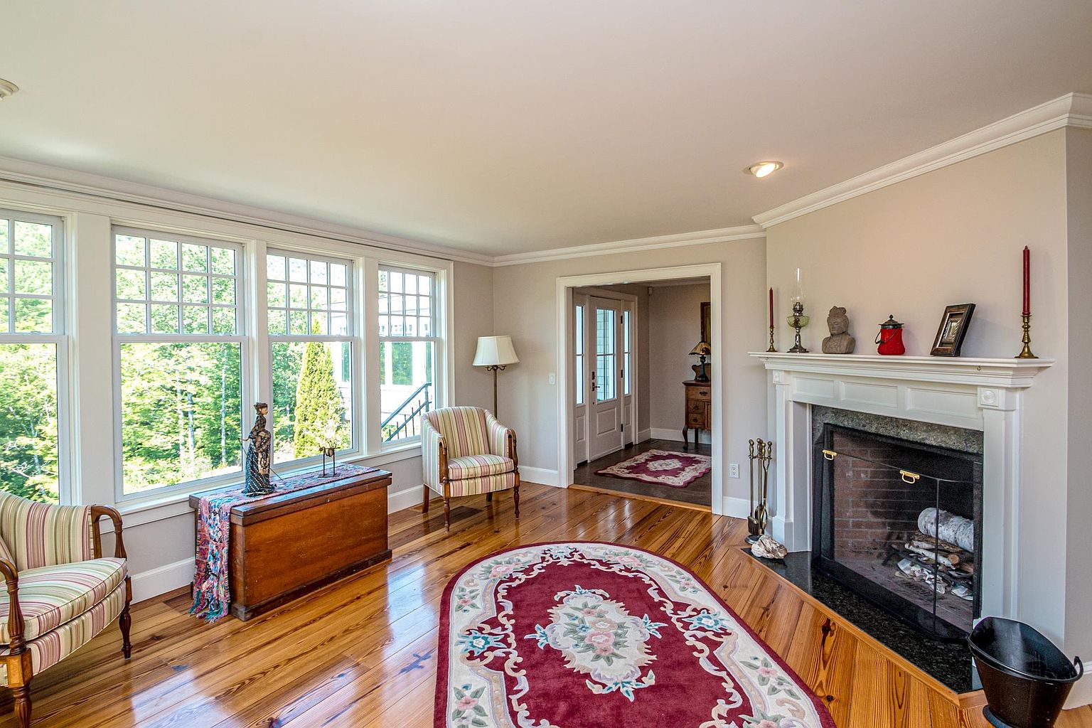 This inviting living room features warm, polished wood flooring and a classic fireplace with a white mantel as a central focal point. Large windows allow ample natural light to fill the space, highlighting the two striped armchairs and the decorative oval rug centered on the floor. The room flows seamlessly into an adjacent hallway, creating an open and airy atmosphere with a traditional, comfortable aesthetic.