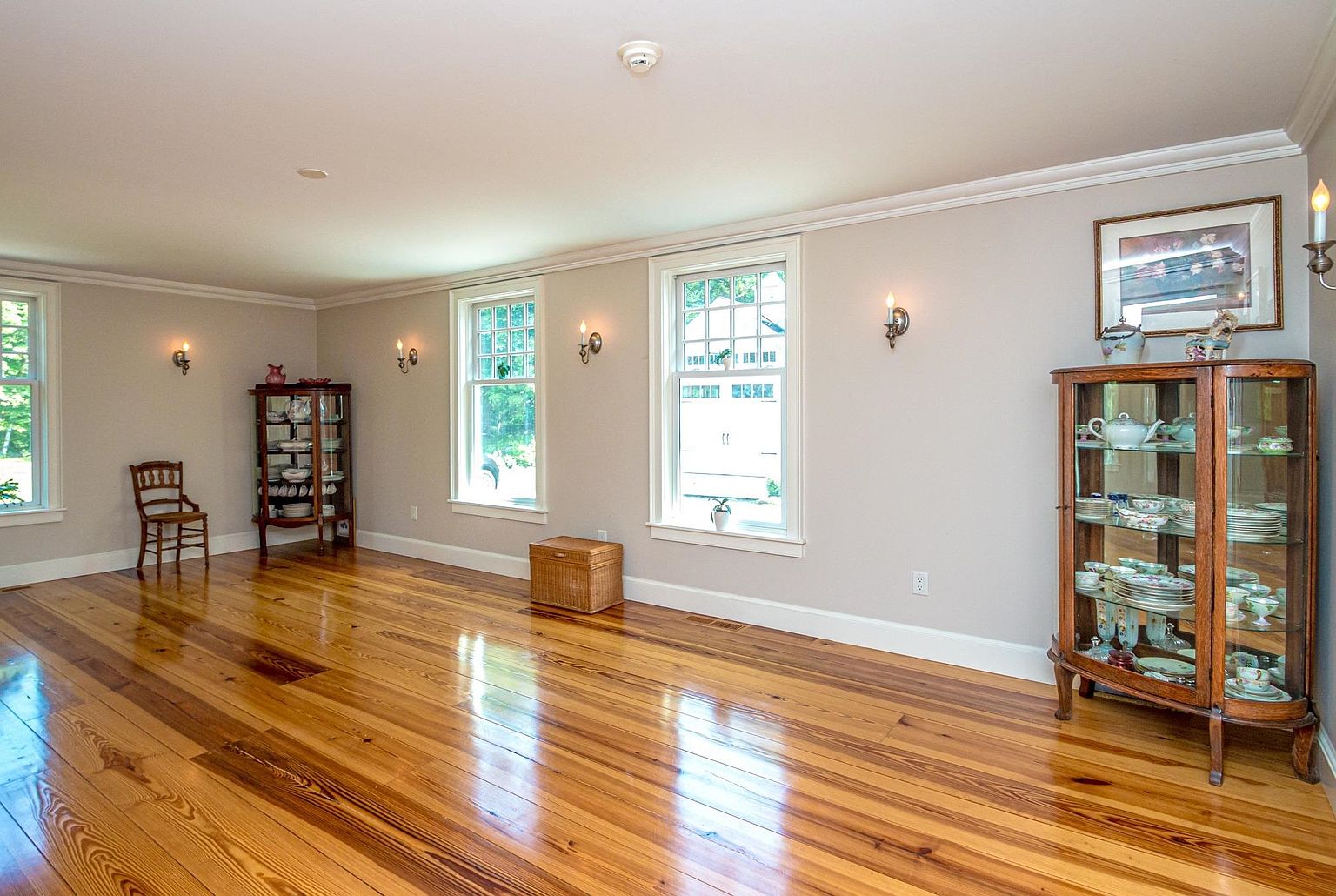 This bright and spacious dining room features polished light wood flooring and neutral-toned walls, creating an airy and inviting atmosphere. The room is furnished with two elegant glass-fronted china cabinets and a single wooden chair, while multiple windows allow for ample natural light. The space is accented by wall-mounted sconces and a framed piece of art, offering a classic and refined aesthetic.