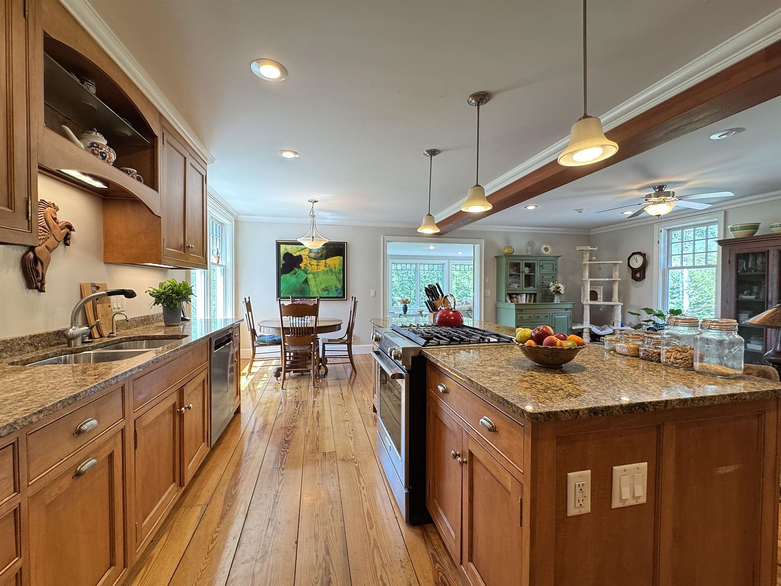 This spacious, warm-toned kitchen features medium-toned wood cabinetry, granite countertops, and a large central island with a gas cooktop. The room flows seamlessly into a dining area with a round table and a sun-drenched breakfast nook beyond, all tied together by polished wood flooring. Pendant lighting hangs over the island, while a large piece of abstract art adds a vibrant focal point to the dining space.