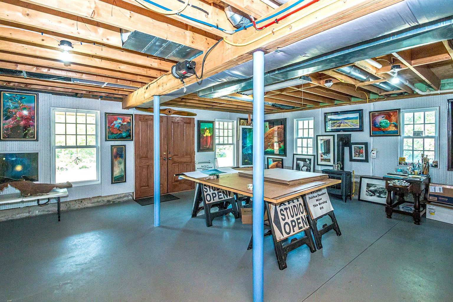 This spacious basement studio features an open layout with exposed ceiling joists and painted concrete floors, creating a functional workspace for artists. The room is equipped with a large central work table, multiple windows that provide natural light, and pegboard walls adorned with various framed artworks. A small wood-burning stove sits in the corner, adding a cozy element to this versatile, industrial-style creative area.