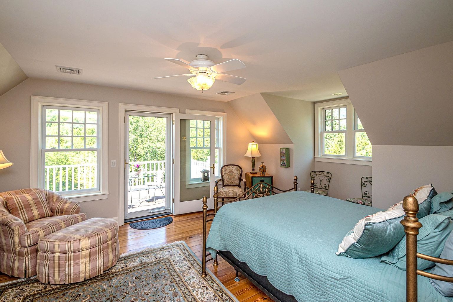 This cozy bedroom features a metal-framed bed with a light blue quilt and a matching plaid armchair with an ottoman. The room is illuminated by a ceiling fan and a bedside lamp, with natural light streaming in from multiple windows and a glass door that opens onto a private balcony. The space is finished with warm wood flooring and a patterned area rug, creating a comfortable and inviting atmosphere.
