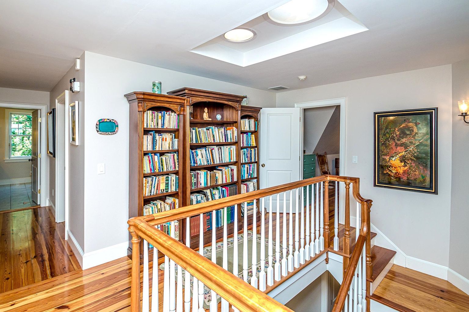 This image captures a bright landing area featuring a large, multi-section wooden bookshelf filled with books. A wooden staircase with white balusters leads down, while a skylight above provides natural illumination to the space. The hallway is finished with warm-toned wood flooring and neutral walls, accented by a framed piece of abstract art.