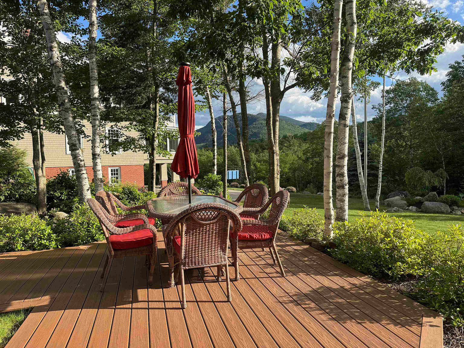 This inviting outdoor living space features a composite deck furnished with a wicker dining set and a red umbrella, perfect for al fresco dining. The deck overlooks a lush, green lawn surrounded by mature trees and offers a scenic view of distant mountains. The setting provides a peaceful, private atmosphere ideal for relaxation and entertaining.