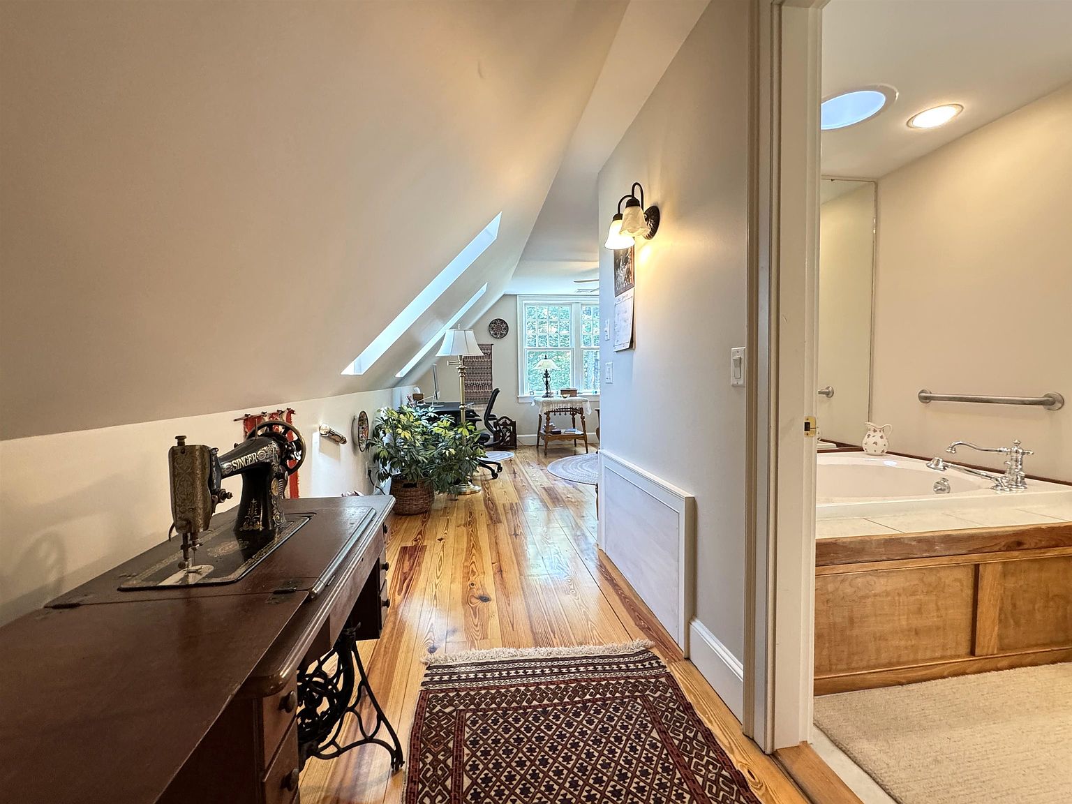 This attic-level hallway features warm-toned wood flooring and a sloped ceiling with skylights that flood the space with natural light. To the left, a vintage Singer sewing machine table serves as a decorative console, while a doorway on the right leads into a bathroom with a soaking tub. The perspective looks down the length of the hall toward a bright window at the far end, creating an inviting and cozy atmosphere.