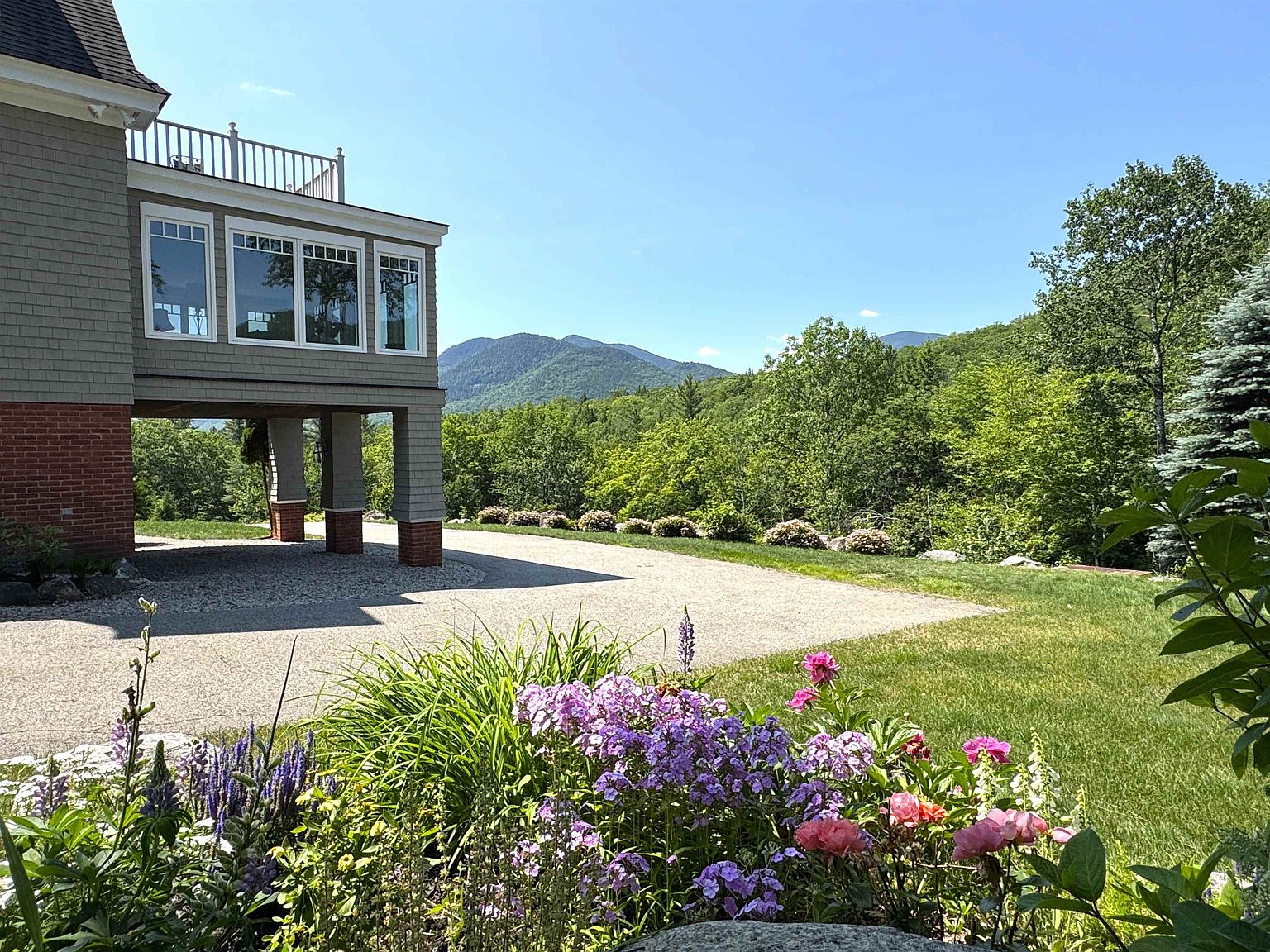 This exterior view captures the rear of a multi-story home featuring a raised deck and a covered lower-level parking area supported by brick-clad pillars. The house is surrounded by a lush, vibrant garden in the foreground and a expansive gravel driveway, all set against a backdrop of rolling green mountains under a clear blue sky. The perspective highlights the seamless transition between the home's architecture and the natural landscape, emphasizing a peaceful, scenic setting.
