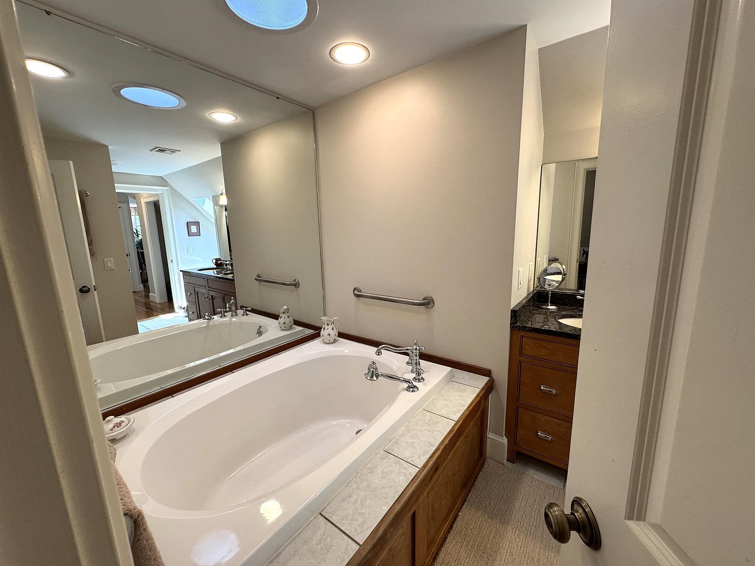 This primary bathroom features a large soaking tub set into a tiled deck with a wood-paneled base, complemented by a wall-mounted grab bar for accessibility. A expansive mirror reflects the room, including a vanity with a dark stone countertop and a separate sink area visible in the background. The space is illuminated by recessed lighting and includes neutral wall tones, creating a functional and traditional aesthetic.