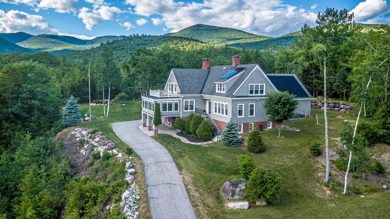 This elevated aerial perspective captures a multi-level shingle-style home nestled against a lush, mountainous backdrop. The property features a paved driveway leading to the residence, which is accented by a wrap-around deck, multiple gables, and a prominent stone chimney. The surrounding landscape is characterized by dense forest, manicured lawn areas, and natural rock outcroppings, creating a serene and secluded atmosphere.