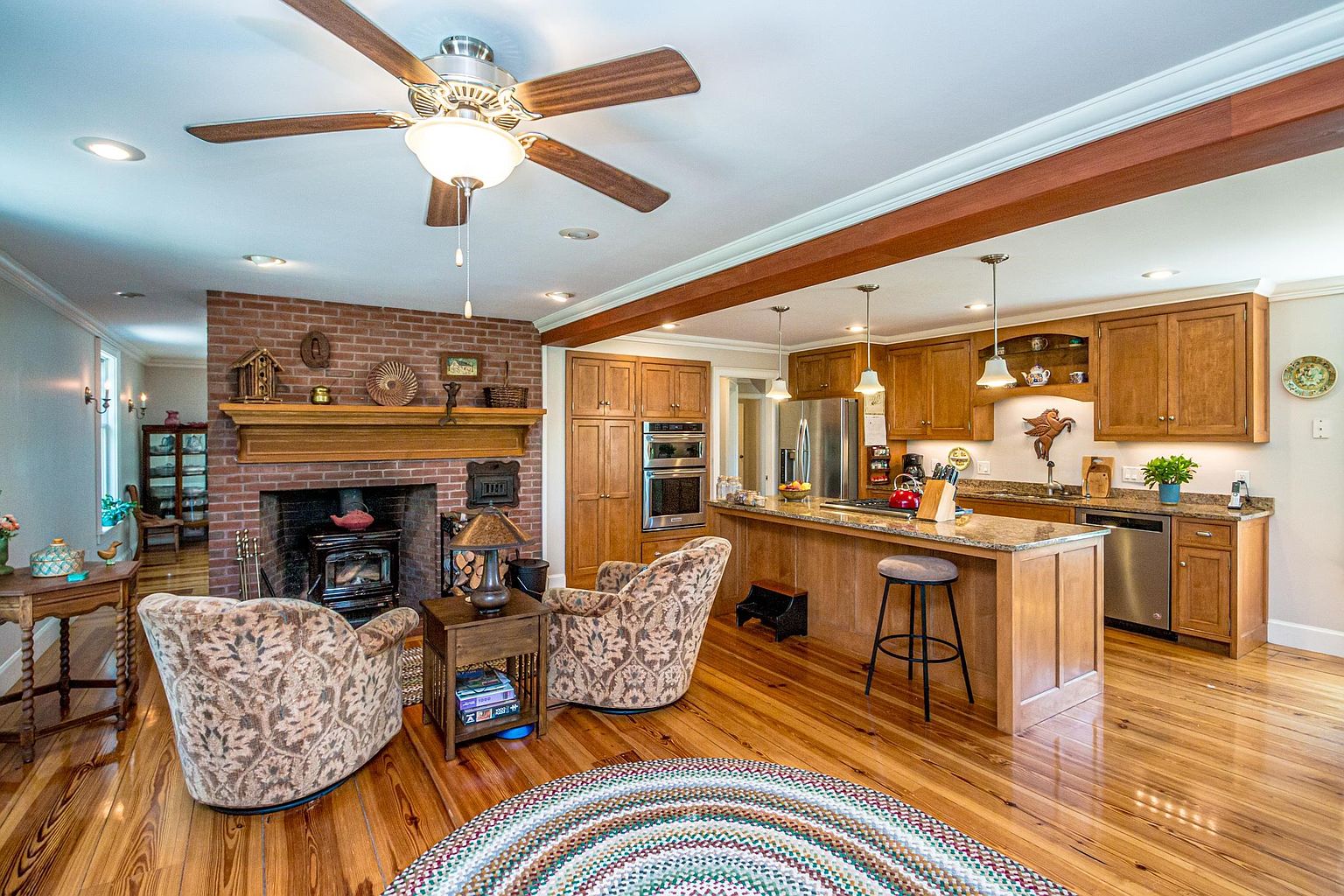 This open-concept space seamlessly blends a cozy living area with a functional kitchen, featuring warm wood cabinetry and polished wood flooring throughout. A prominent brick chimney breast serves as a focal point for the living area, which includes two patterned armchairs and a central rug, while the kitchen offers a large island with bar seating and stainless steel appliances. The room is illuminated by a combination of recessed lighting, pendant lights over the island, and a ceiling fan, creating a welcoming and cohesive atmosphere.