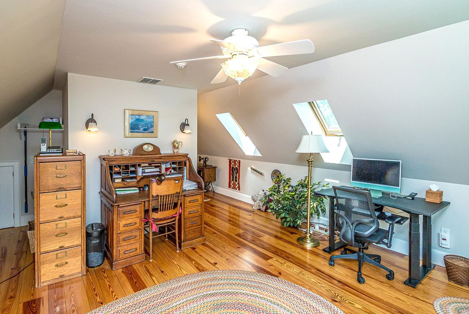 This spacious home office is situated in an attic space with sloped ceilings and skylights that provide ample natural light. The room features warm-toned wood flooring, a traditional roll-top desk, and a modern standing desk setup, creating a blend of classic and contemporary workspaces. A large, colorful circular rug anchors the center of the room, while a ceiling fan and wall-mounted lamps provide additional lighting.