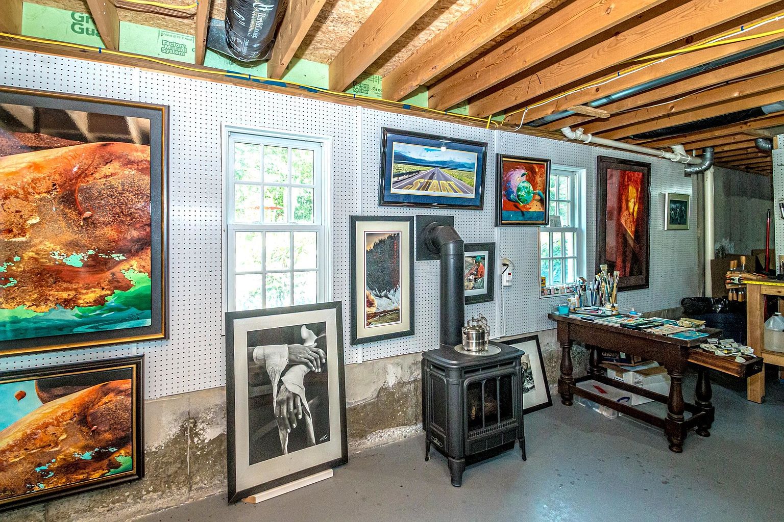 This basement space functions as an art studio, featuring pegboard walls adorned with various framed artworks and a cast-iron wood-burning stove. A wooden work table sits to the right, cluttered with art supplies, while the unfinished ceiling exposes wooden joists and utility pipes. The room has a utilitarian aesthetic with concrete flooring and natural light filtering through two windows.
