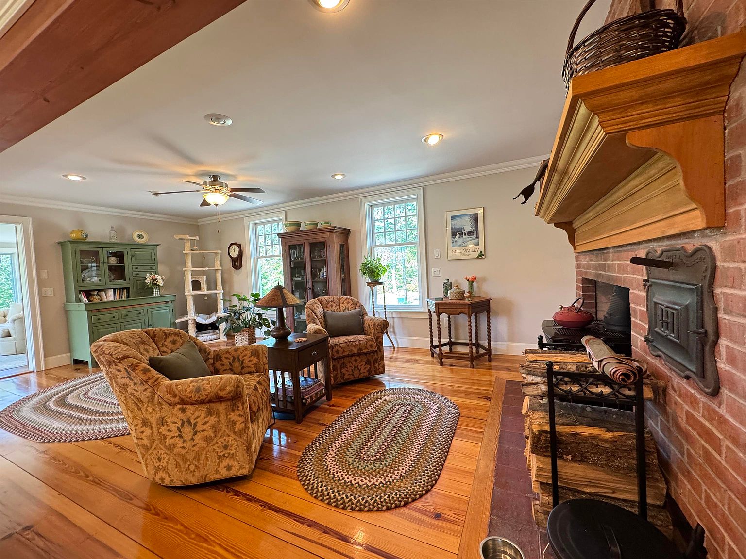 This cozy living room features warm wood flooring, a brick chimney breast with a wood-burning stove, and a prominent wooden mantel. The space is furnished with two patterned armchairs, a small side table, and a green hutch, all arranged around braided area rugs. Large windows allow natural light to fill the room, creating a welcoming and traditional atmosphere.