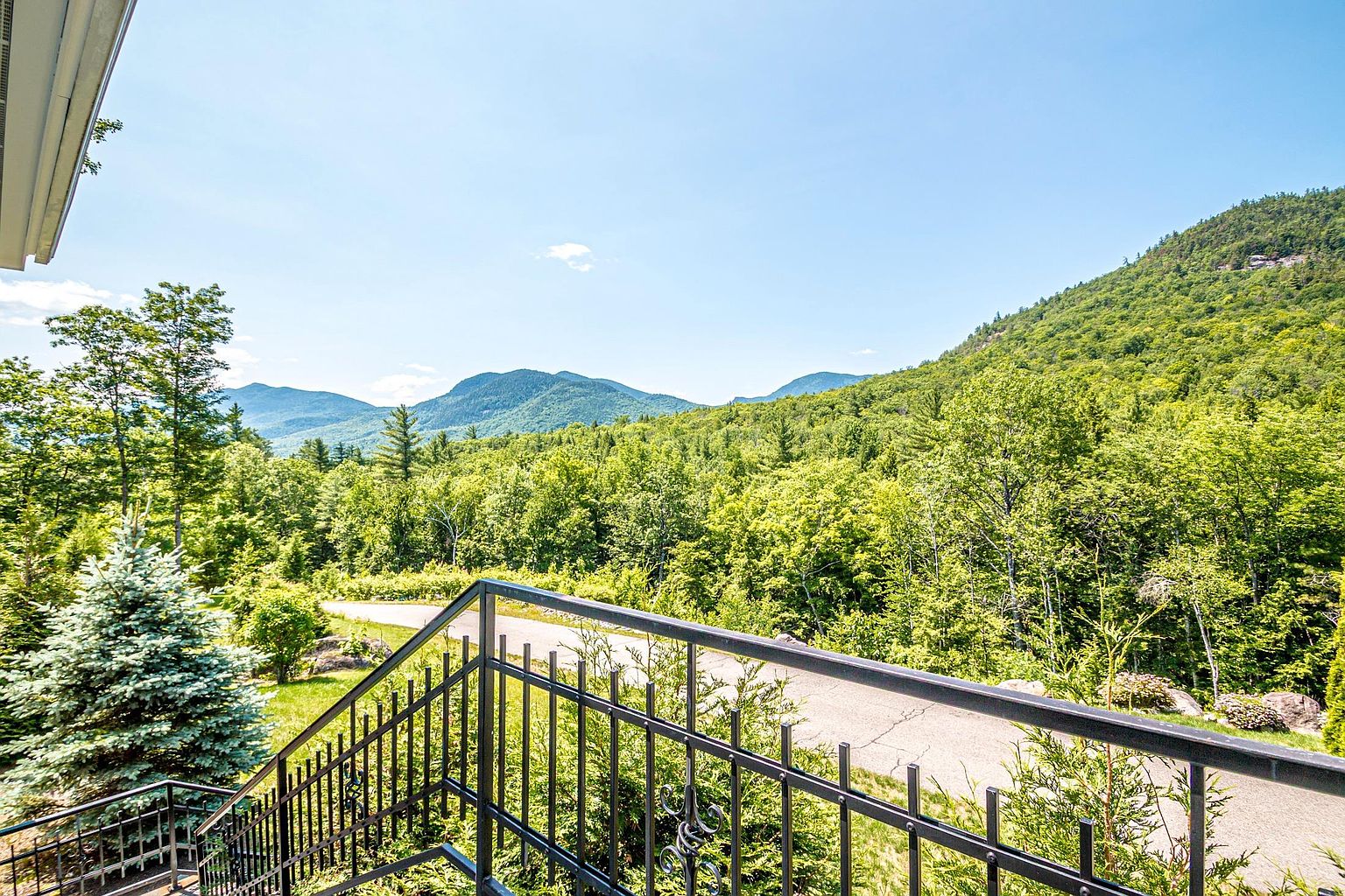 This elevated balcony offers a sweeping, panoramic view of a lush, green mountain range under a clear blue sky. A black metal railing frames the foreground, leading the eye toward the dense forest canopy and the winding driveway below. The perspective is from an upper level, emphasizing the serene and private nature of the property's natural surroundings.