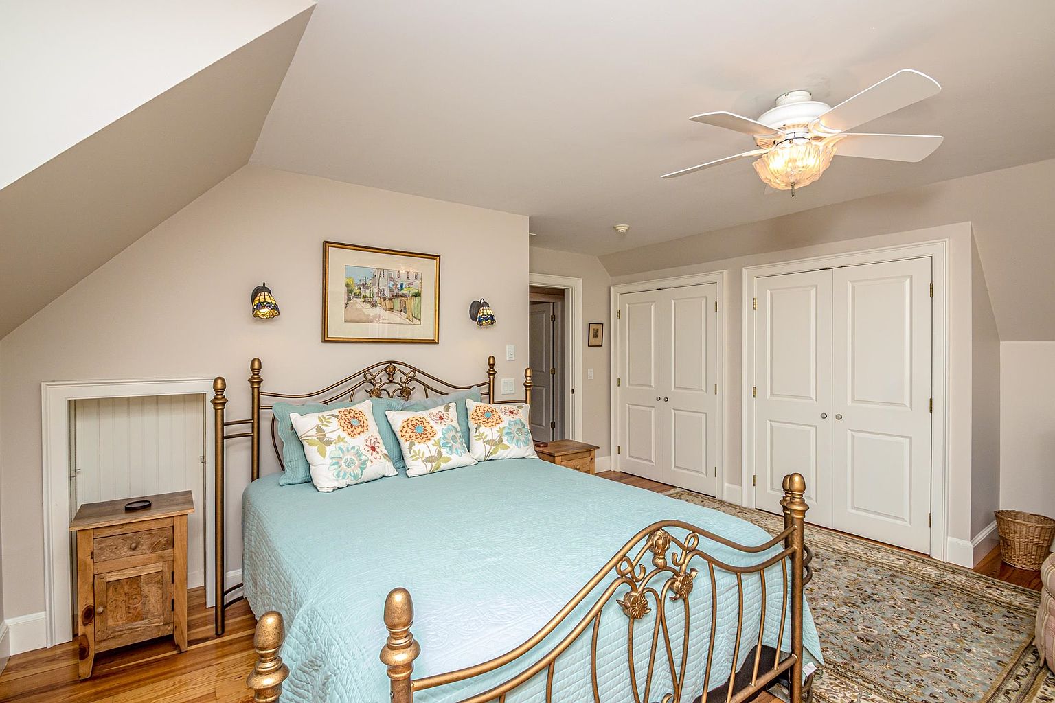 This cozy bedroom features a metal-framed bed with a light blue quilt and floral accent pillows, set against a neutral wall color. The room includes a small wooden nightstand, a framed landscape print, and two wall-mounted sconces, all situated under a sloped ceiling. A patterned area rug covers the wood flooring, and double doors lead to a closet, creating a warm and inviting atmosphere.