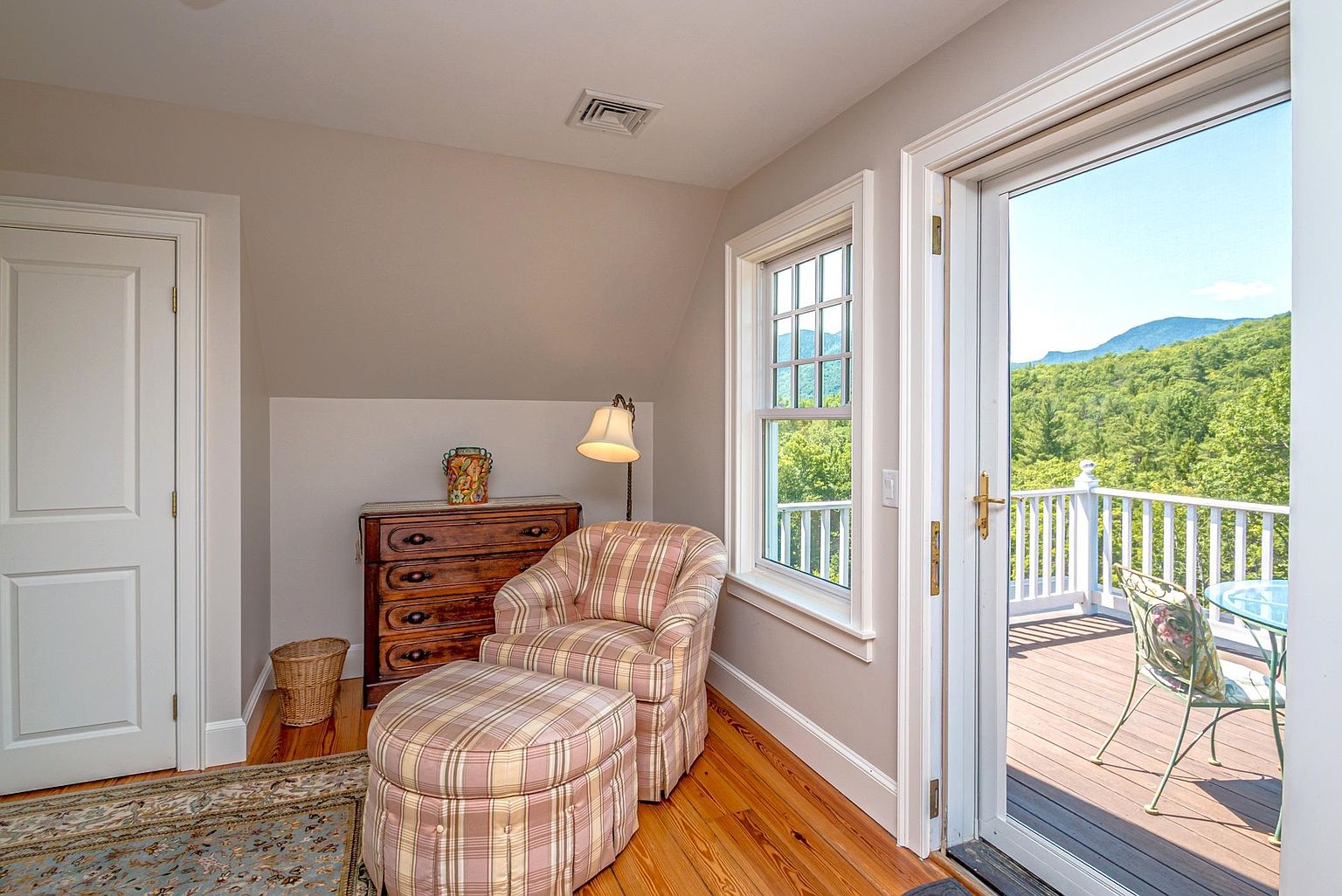 This cozy, light-filled room features warm wood flooring and a comfortable plaid armchair with a matching ottoman positioned near a window. A wooden dresser sits against the wall, topped with a decorative lamp, while a glass door provides direct access to an outdoor deck with scenic mountain views. The space offers a peaceful, inviting atmosphere perfect for a quiet reading nook or guest retreat.
