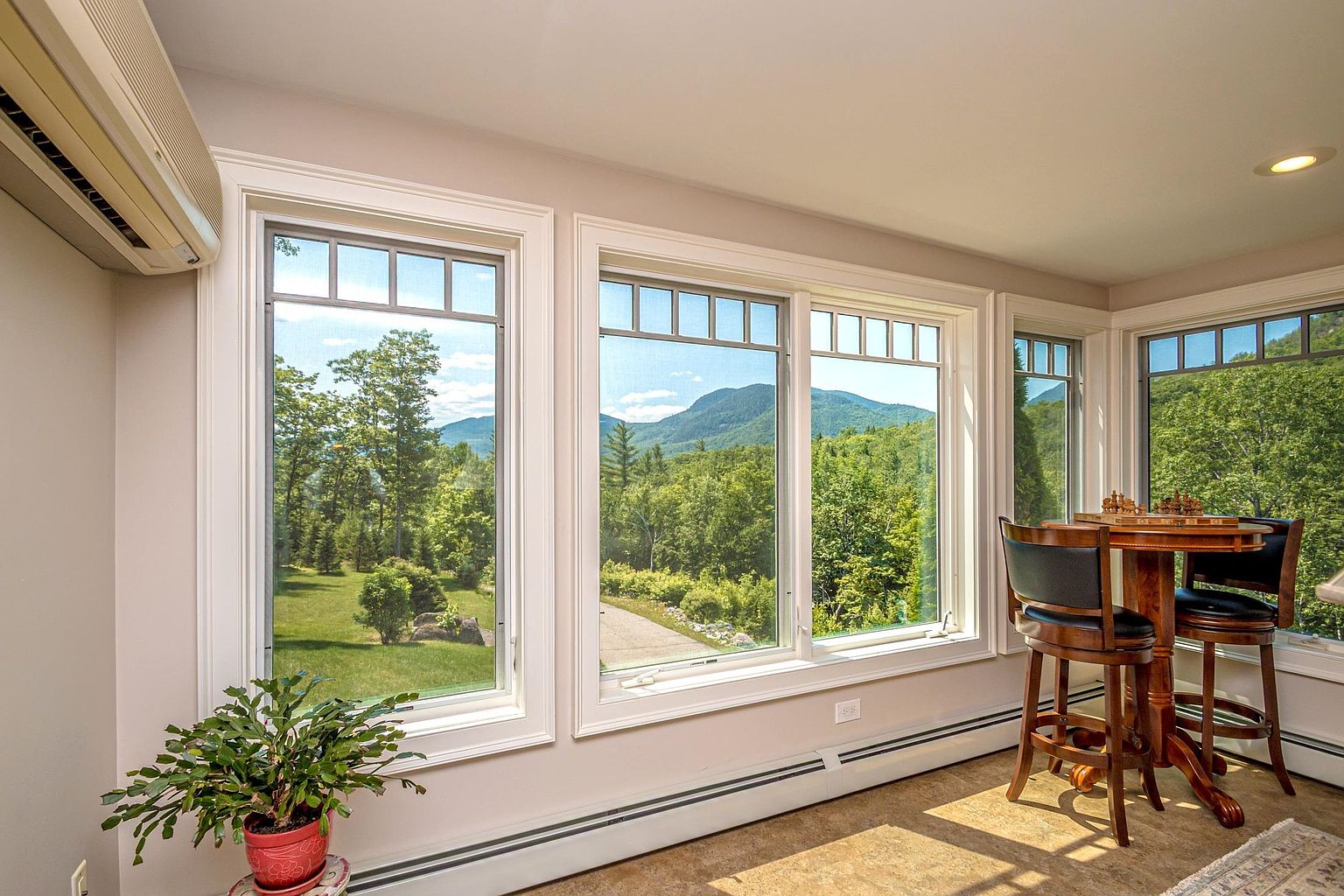 This bright sunroom features a series of large windows that frame expansive views of the surrounding forest and distant mountains. A small wooden bistro table with two chairs sits in the corner, creating a cozy nook for relaxation or games. The space is finished with neutral walls, baseboard heating, and a potted plant, offering a serene and airy atmosphere.