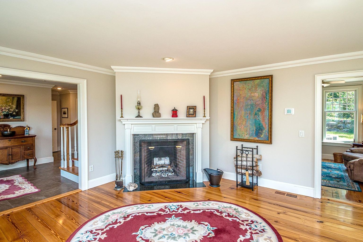 This living room features a central fireplace with a white mantel and a dark stone surround, serving as the focal point of the space. The room is anchored by a large, circular patterned rug on polished wood floors, with a tall, colorful abstract painting displayed on the adjacent wall. The perspective captures the room's open layout, showing transitions into a staircase area on the left and a sunlit room on the right.