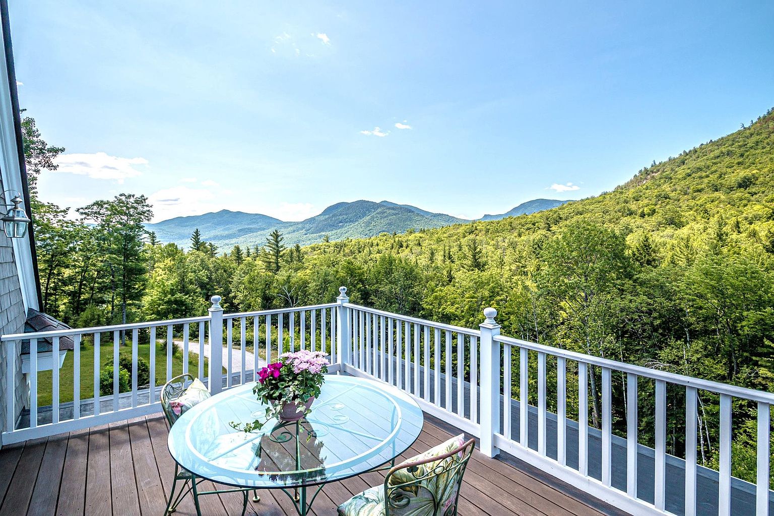 This elevated wooden deck offers a panoramic view of lush, green mountain ranges under a clear blue sky. The space is furnished with a round glass-topped table and two patterned chairs, accented by a potted plant with vibrant pink flowers. A white railing encloses the deck, providing a secure vantage point to enjoy the expansive natural scenery.