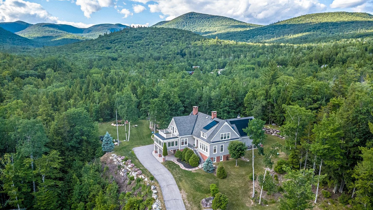 This high-angle aerial shot captures a multi-level residence nestled within a lush, forested mountain landscape. The home features a complex roofline with solar panels, a wrap-around driveway, and a prominent balcony overlooking the surrounding greenery. The expansive, scenic backdrop of rolling hills and dense woods emphasizes the property's secluded and tranquil setting.